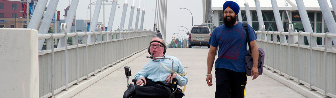 A person sits in a wheelchair and another person stands to his left on a bridge